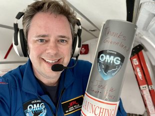 Photo of JPL researcher Josh Willis aboard a small aircraft wearing a flightsuit and holding a cylindrical launching tube used in the OMG mission.