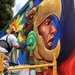 A man on scaffolding is painting a mural of a face wearing a bird mask in profile.