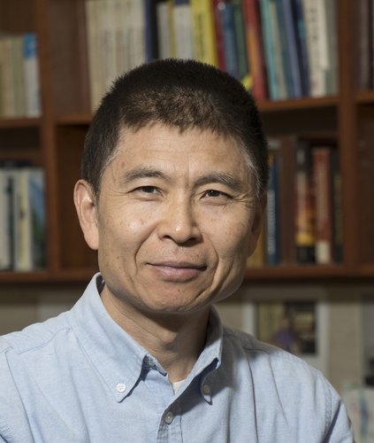 A portrait of Zhen-Gang Wang. He wears a dress shirt and appears in front of shelves of books.