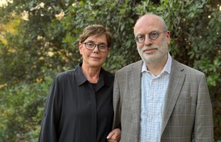 Diana Kormos-Buchwald and Michael R. Gordin stand next to one another arm-in-arm. Kormos-Buchwald wears a long black shirt and Gordin wears a tan plaid sportsjacket.
