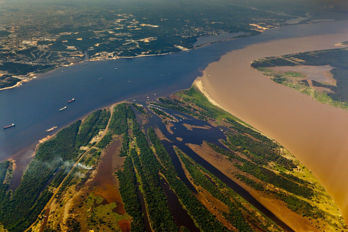 An aerial photo of two rivers joining into one. The flow from one river is dark while the other is muddy. They flow together in the larger, joined river, but remained unmixed.