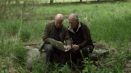 two men sit in a field looking at a computer together