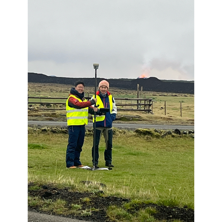 Two people in green reflective vests stand on grass in the foreground while far in the background a red glow of lava can be seen on the horizon