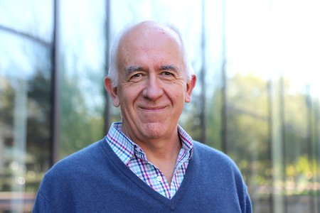 Pietro Perona wearing a collared shirt and blue sweater standing in front of a windowed building