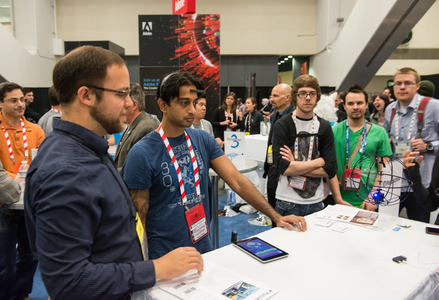 A tech exhibition attendee demonstrates a forehead device near a tablet and drone. Observers in casual clothes surround booths, including one labeled Adobe.