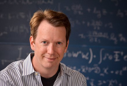 Headshot of Sean Carroll in striped shirt in front of a blackboard with equations