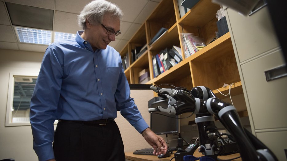Richard Andersen, Caltech's James G. Boswell Professor of Neuroscience, wearing a blue shirt, stands in his office interacting with a robotic arm on a desk. Shelves filled with books and binders are visible in the background.
