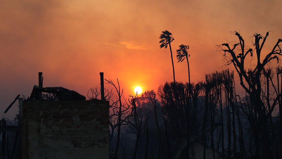 Burned trees and buildings in the foreground and a bright orange sun with a smoky sky in the background