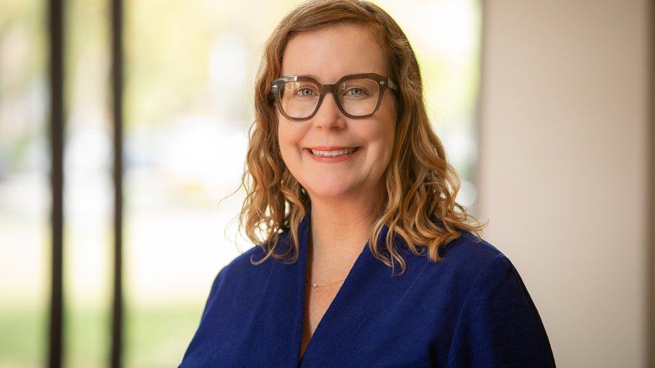 Professor Susanne Hall, wearing glasses and a dark blue top, smiles in front of a softly blurred background with large windows and greenery visible outside.