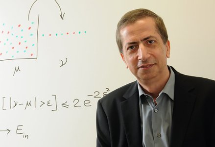Portrait of Yaser Abu-Mostafa, man in suit jacket sitting in front of a whiteboard with equations