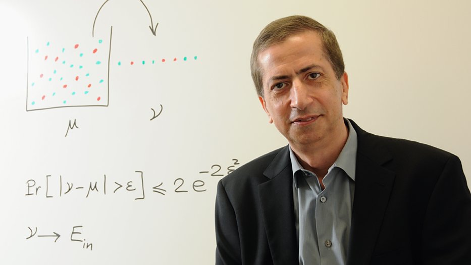 Portrait of Yaser Abu-Mostafa, man in suit jacket sitting in front of a whiteboard with equations