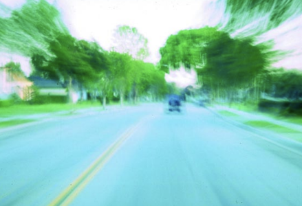 A motion-blurred view of a suburban street seen from a moving vehicle. The road extends into the distance, lined with trees and houses, creating a sense of speed and disorientation.