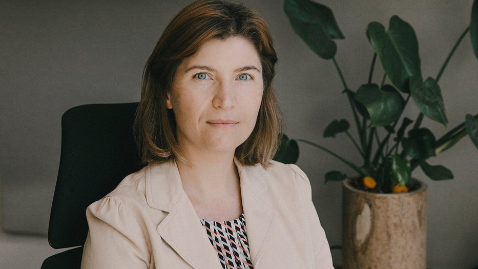 A portrait of Viviana Gradinaru, seated in an office chair, in a light-colored blazer and patterned blouse. A potted plant with broad green leaves is visible in the background.