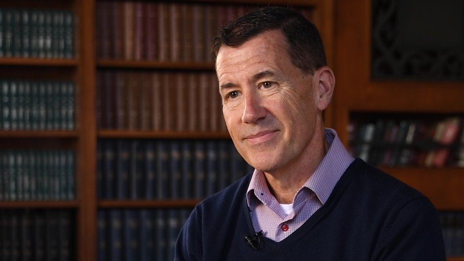 person in a sweater and collared shirt in front of a shelf full of books