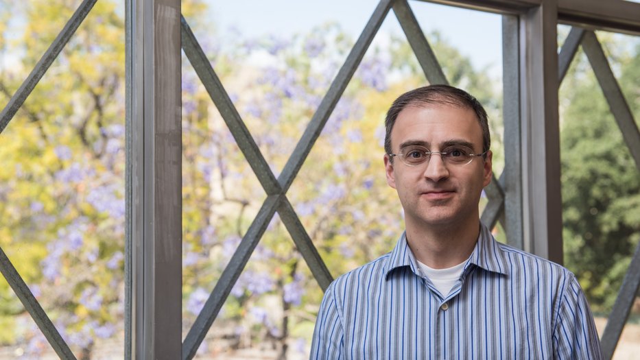 Professor David Prober, wearing a striped blue and white shirt, stands in front of a large window with a geometric frame. Behind him, trees with green and purple foliage are visible on a sunny day.
