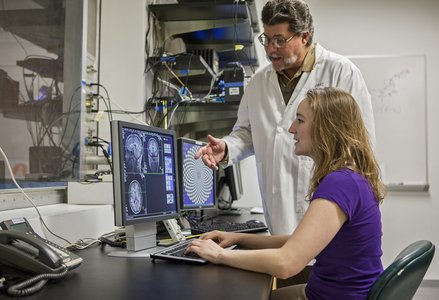 A neuroscientist in a lab coat and a researcher are collaborating on work at a computer displaying brain scan images. The background includes various lab equipment and wires, illustrating a high-tech neuroscience lab setting.