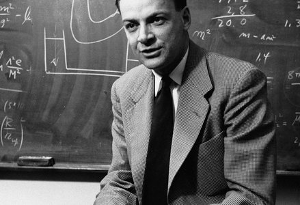 Richard Feynman sitting on a desk in front of a chalkboard with equations
