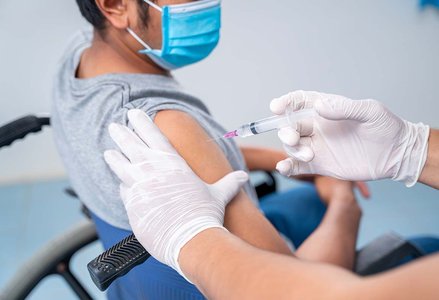 person in a wheelchair receiving an injection in the arm wearing surgical mask