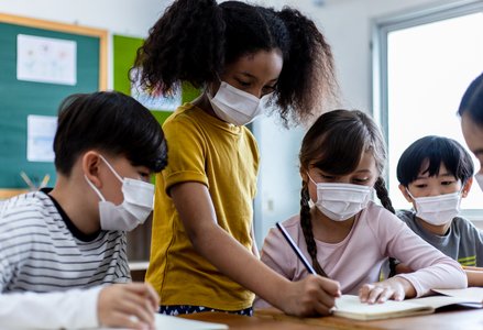 Four elementary school-aged students huddle over a piece of paper wearing surgical masks