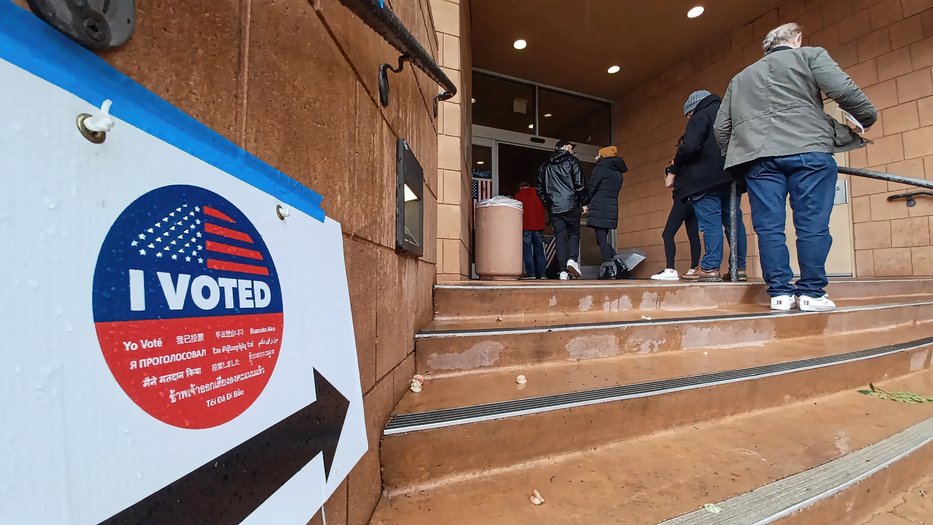 People stand in line outside a voting center. A sign with 'I VOTED' and an American flag design is on the left, featuring translations in multiple languages. An arrow points toward the entrance, guiding voters to the building.