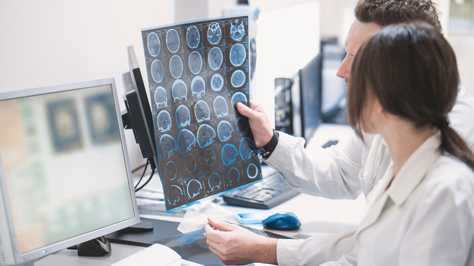 Two medical professionals analyze brain scans on transparent film in a clinical setting, focusing on cross-sections of the brain for research or diagnosis.