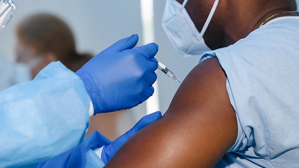 Person in personal protective equipment administers a shot in the arm of a man wearing a kn95 mask