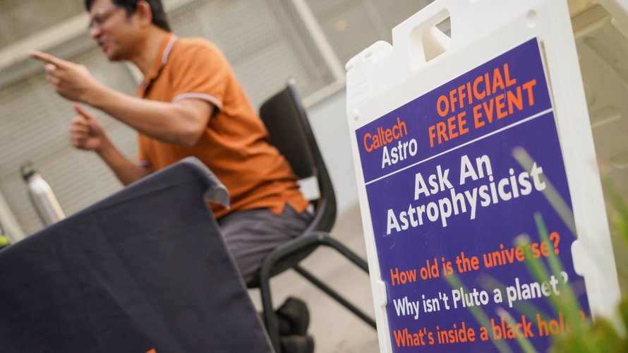 A man talks at a table behind a sign that reads "Ask an Astrophysicist."