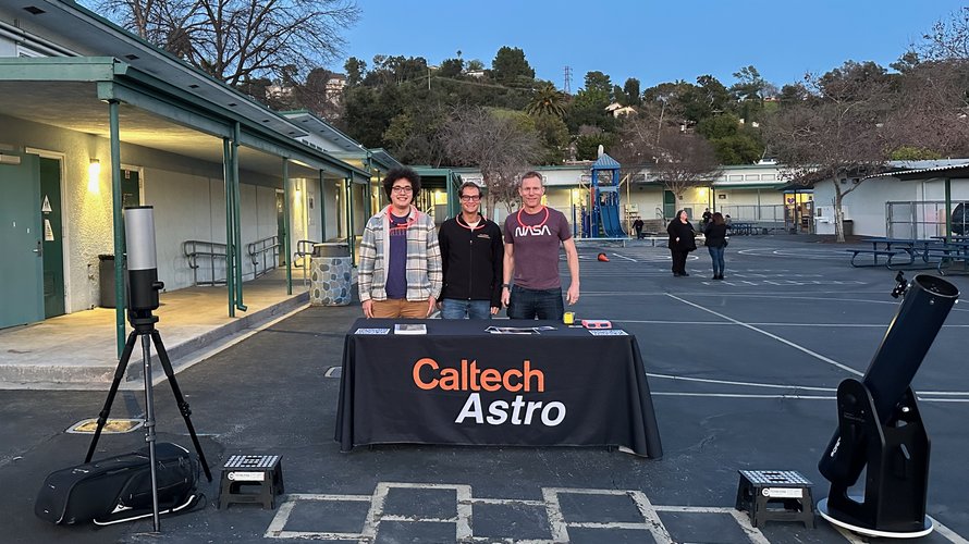Three astronomers standing behind an outdoor table that says "Caltech Astro," flanked by two telescopes.