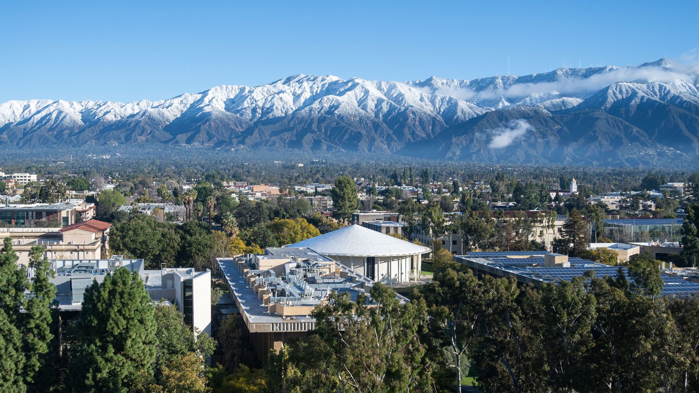Beckman Auditorium from above surrounded by trees with the the snow-capped San Gabriel Mountains in the background.