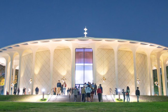 A crowd of all ages enters Beckman Auditorium at night.
