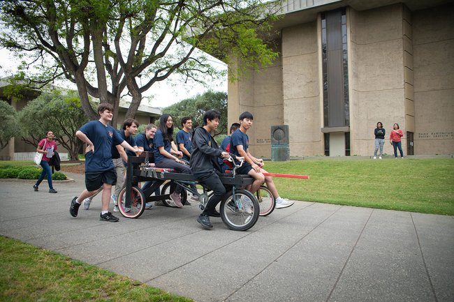Eight students in matching shirts riding a makeshift vehicle on Ditch Day.