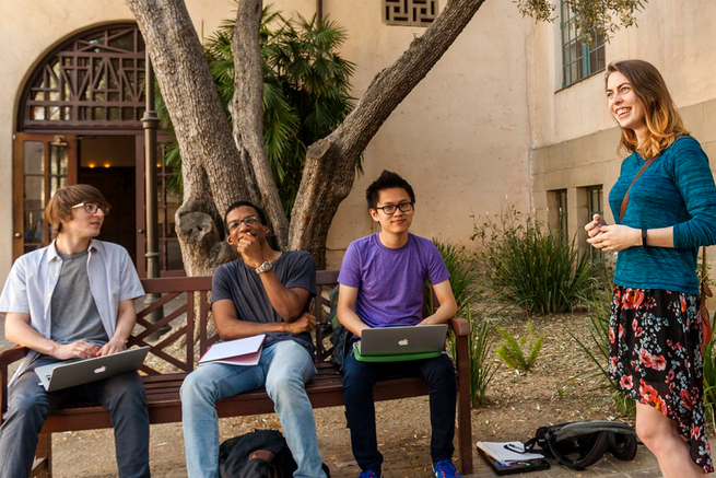 Humanities students sitting on a bench under a tree talking with laptops open.
